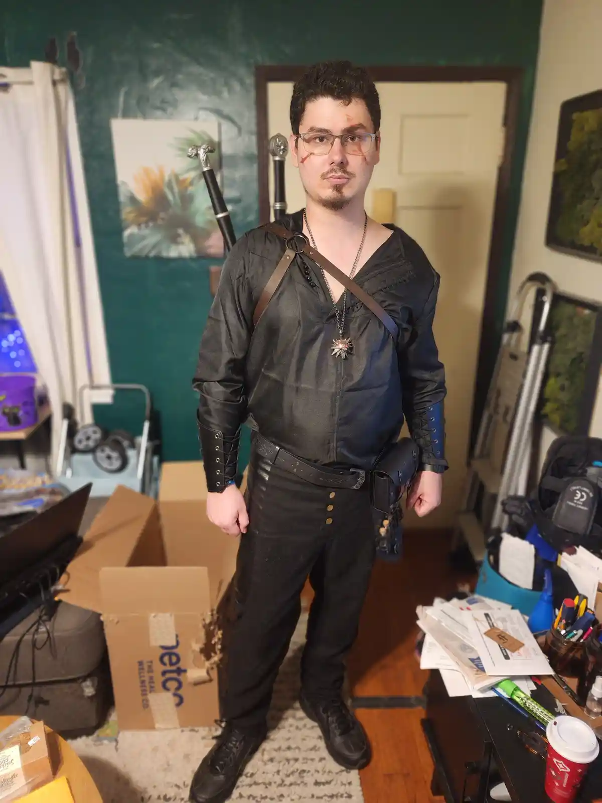 A man in a detailed Geralt of Rivia cosplay from The Witcher, complete with swords on his back, a silver star pendant, and…