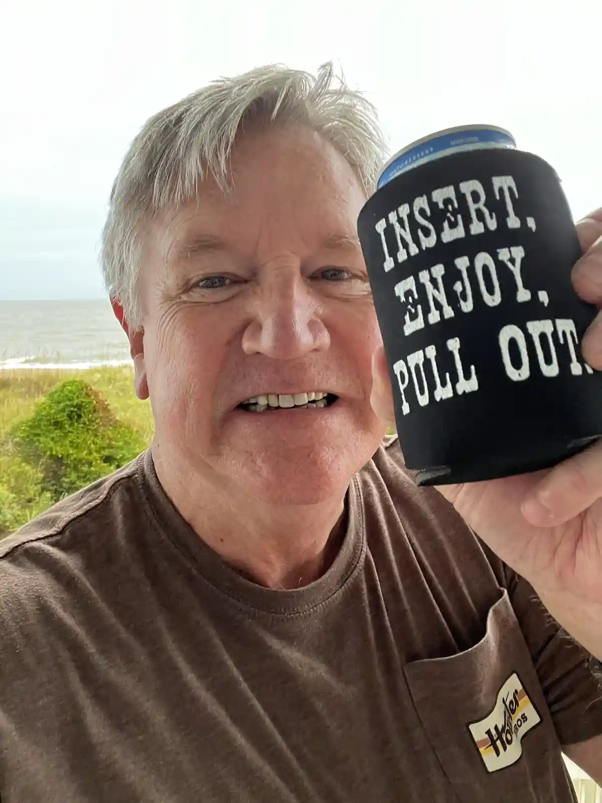 A smiling older man with gray hair holds up a black can cooler with white text reading 'INSERT, ENJOY, PULL OUT' over a…
