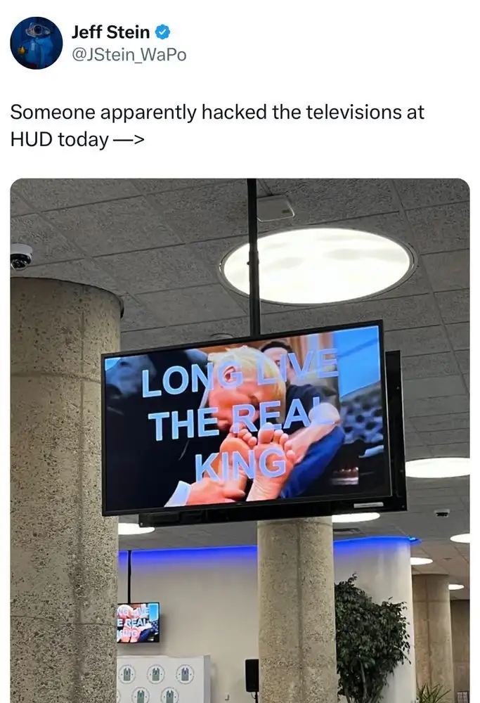 A television screen in a public building displays the text 'LONG LIVE THE REAL KING' over an image of a person holding a…