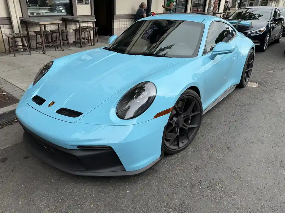 A light blue Porsche 911 GT3 sports car is parked on a city street, showing its front and driver's side. The car has black…