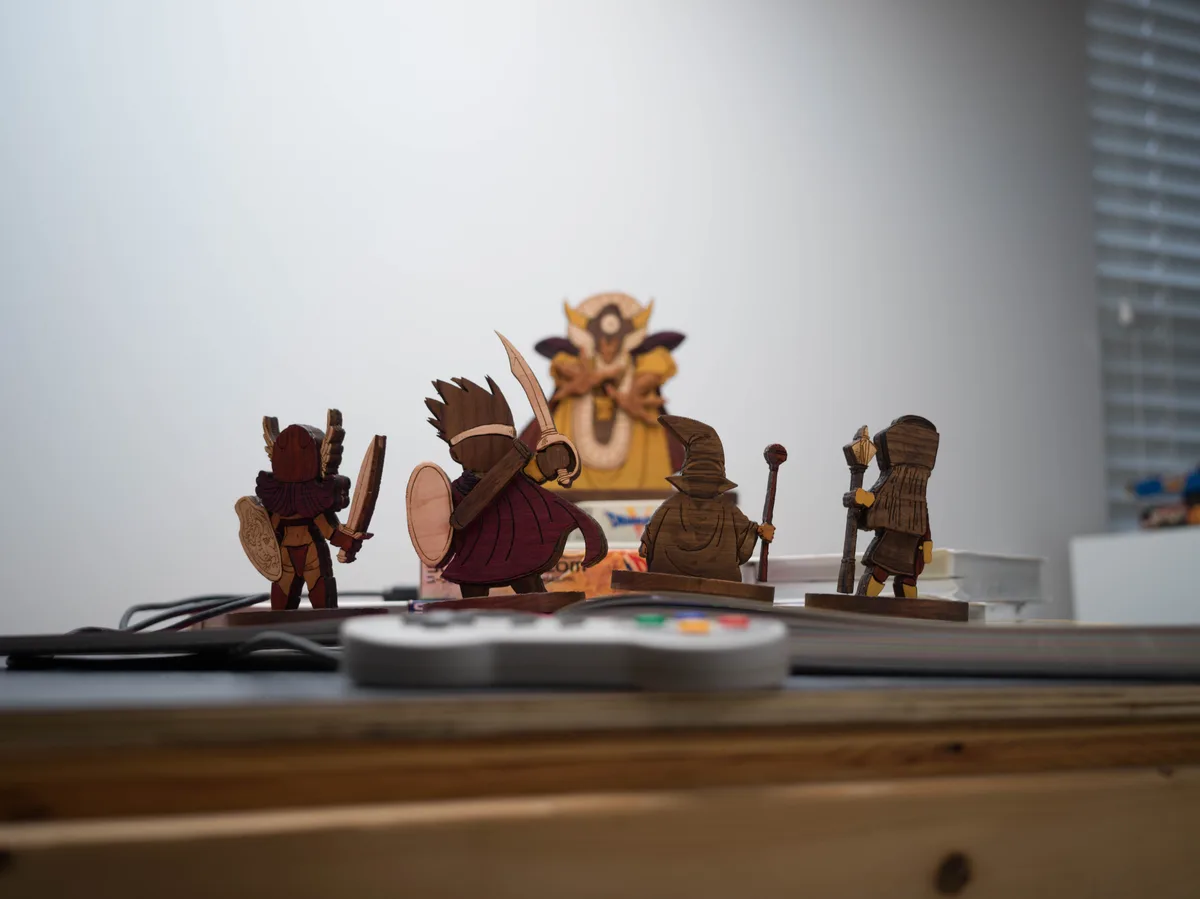 A photo of four wooden fantasy miniatures—a warrior, a swordsman, a wizard, and a cleric—standing on a desk in front of a…