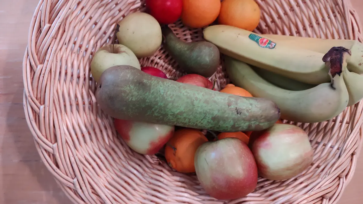 A wicker basket filled with various fruits including apples, oranges, and bananas, but prominently features a long, green…