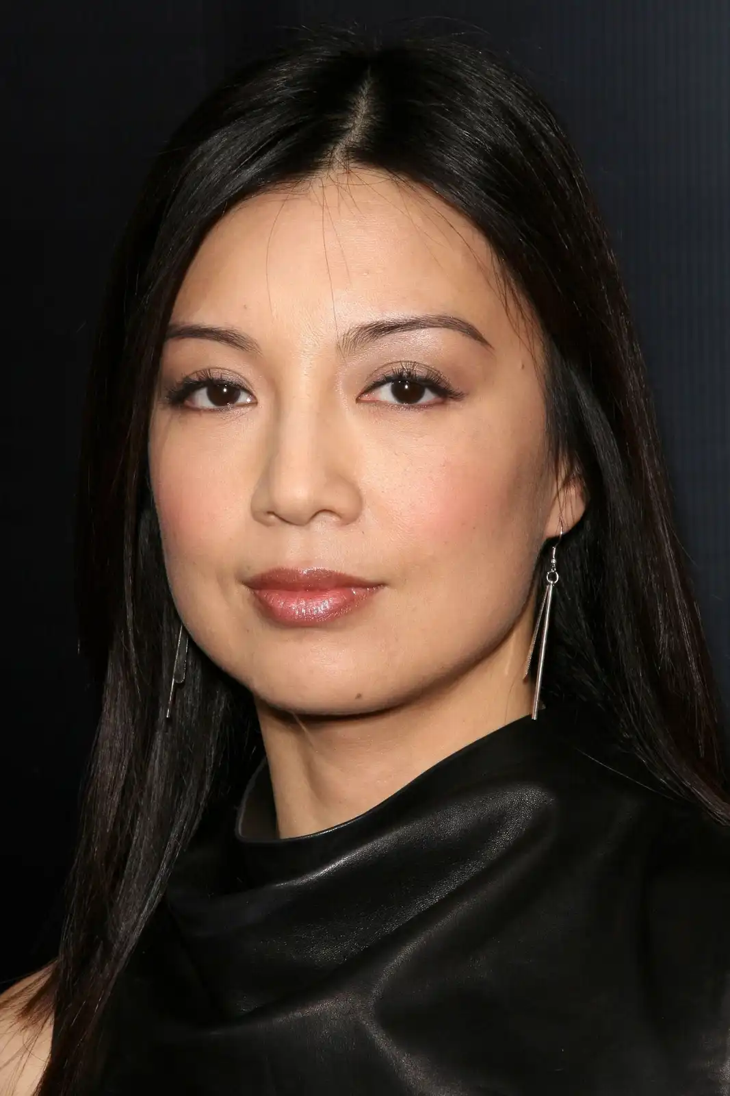 A close-up photo of a woman with long dark hair, wearing a black leather top and silver dangling earrings, looking directly…