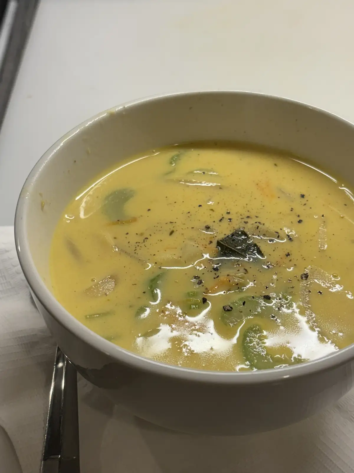 A close-up photo shows a white ceramic bowl filled with creamy yellow soup, garnished with black pepper and a dark leaf…