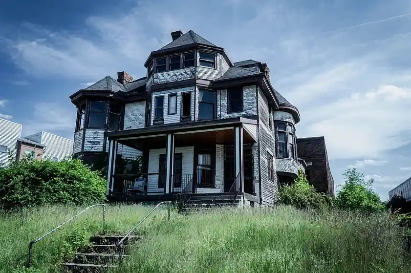 An old, dilapidated Victorian-style house with peeling paint and broken windows stands surrounded by tall, overgrown grass…