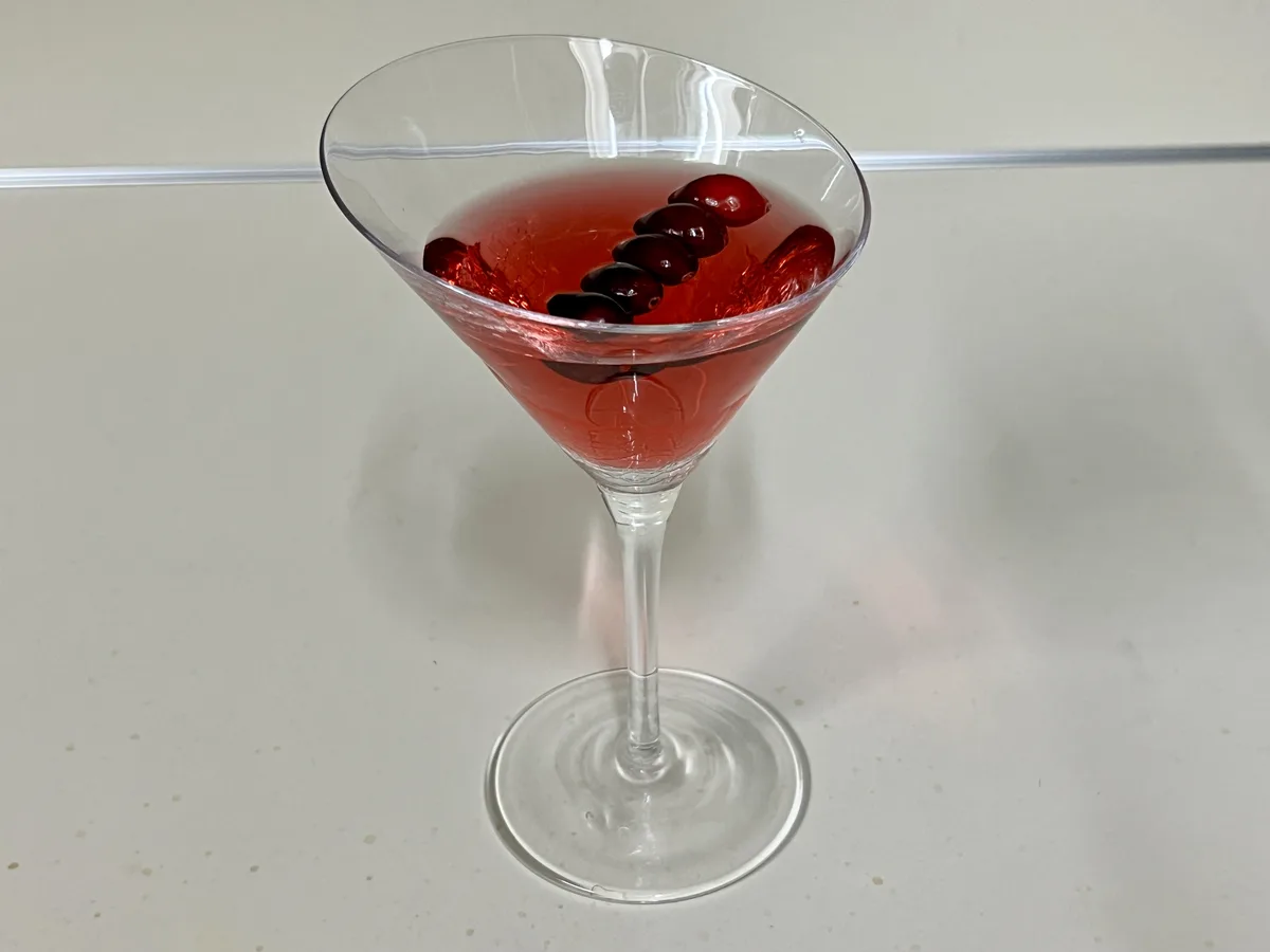 A clear martini glass sits on a light-colored countertop, filled with a red liquid and garnished with several dark red…