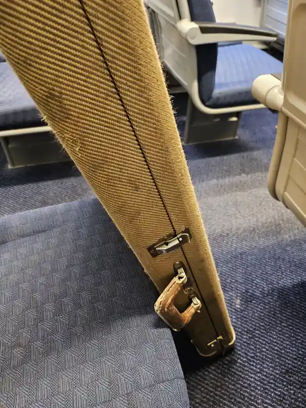 A close-up photo shows a worn, beige, fabric-covered suitcase with a metal latch and a frayed, brown leather handle, resting…