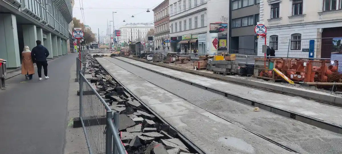 A street view of tram tracks under construction, with broken pavement and construction materials piled along the rails. Two…