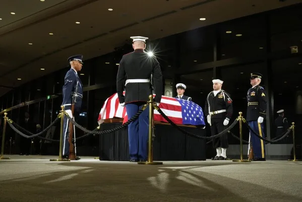 A solemn military funeral scene with five uniformed service members standing at attention around a casket draped with an…
