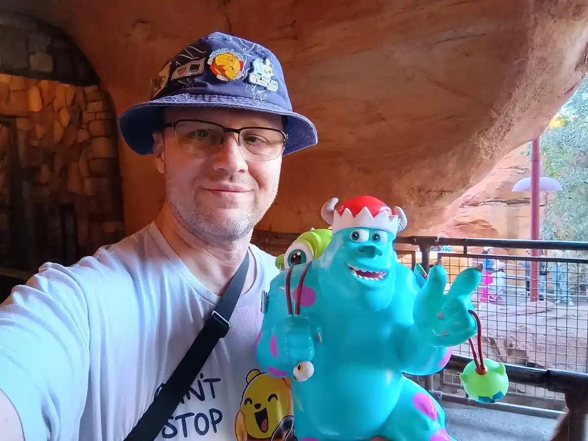 A man wearing glasses and a blue bucket hat with pins takes a selfie next to a large, cheerful blue monster statue wearing a…