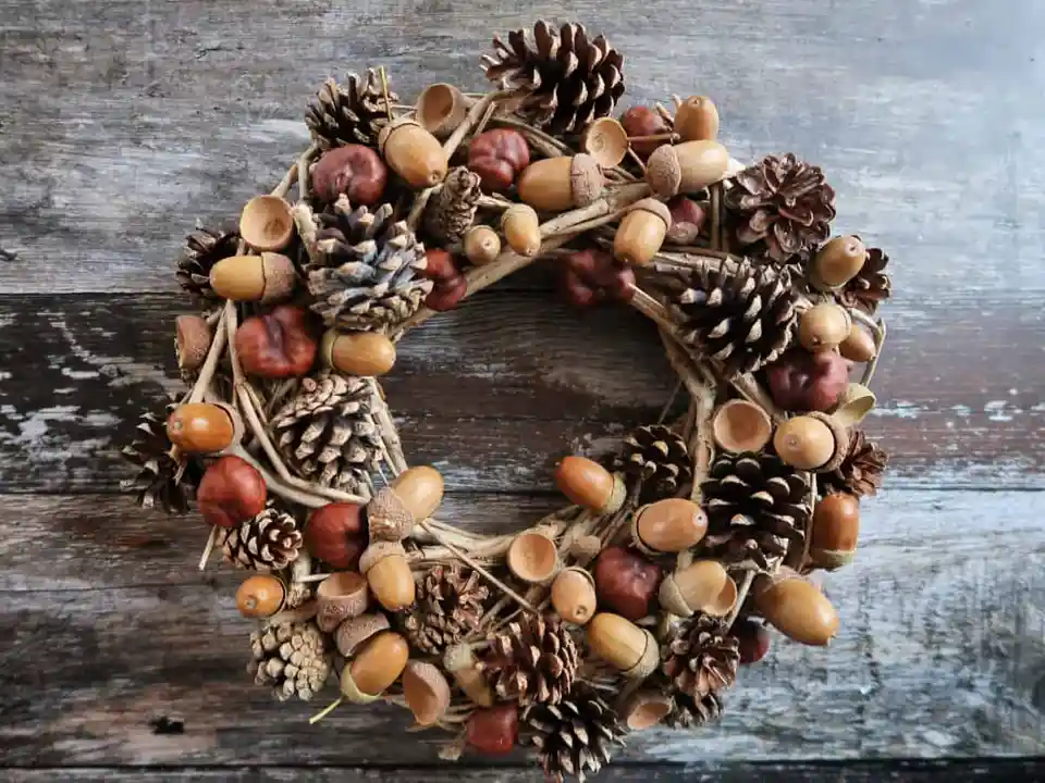 A rustic autumn wreath made of twigs, pinecones, acorns, and chestnuts sits on a weathered wooden surface. Overlaid text…