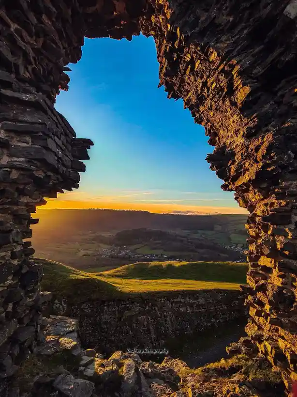 A scenic photograph shows a view through a rough, irregular stone window frame of an ancient ruin, looking out onto a valley…