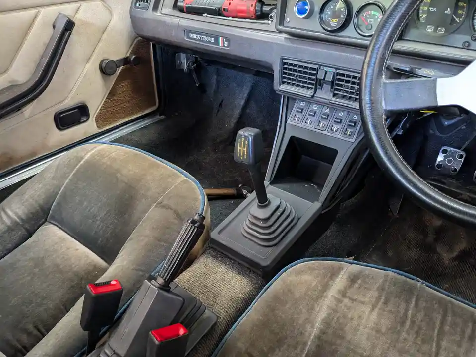 The image shows the interior of a vintage car, likely a Lancia Beta or similar, with worn beige and grey fabric seats, a…