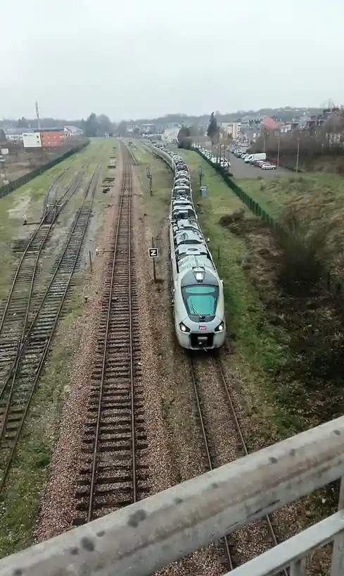 An aerial view shows a long silver SNCF train on a railway track, viewed from a bridge. The train is moving away from the…