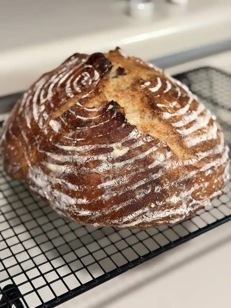 A round, golden-brown loaf of artisan bread with flour dusting and scoring sits on a black wire cooling rack. The overlaid…