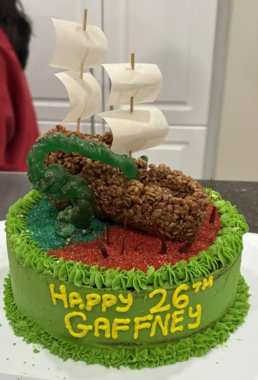 A round green-frosted birthday cake with yellow icing text reading 'Happy 26th Gaffney' is decorated with a rice krispie…