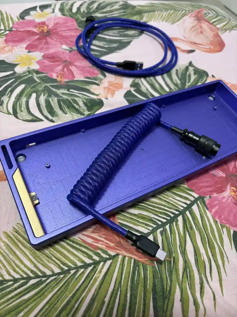 A blue coiled USB-C cable lies on a blue keyboard case, which rests on a tropical-patterned fabric with pink hibiscus…