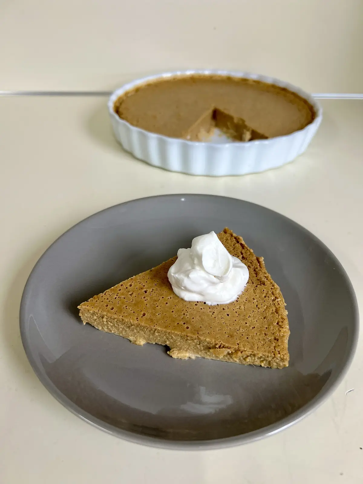 A slice of brown, textured pie topped with whipped cream sits on a gray plate in the foreground, with the rest of the pie in…