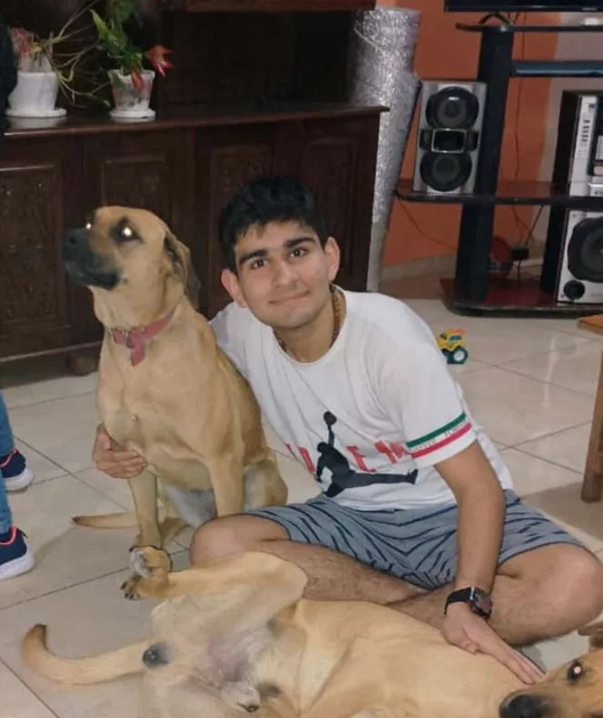 A young man sits on a tiled floor in a living room, smiling at the camera while holding one light-brown dog upright and…