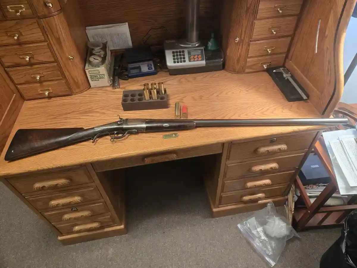 A vintage rifle with a long barrel lies horizontally on a wooden desk surrounded by reloading equipment, brass casings, and…