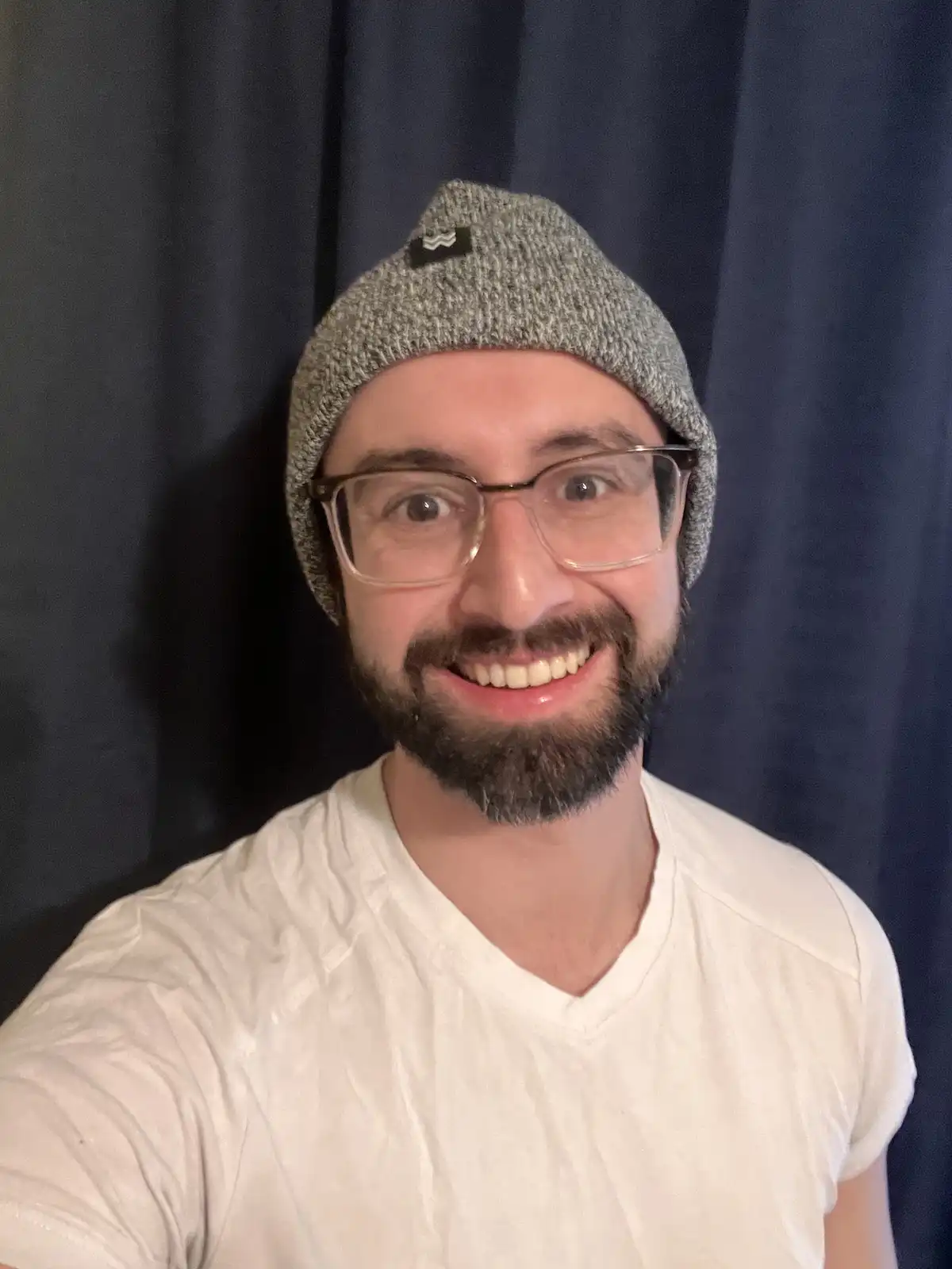 A man with a beard and glasses, wearing a grey knit beanie and a white v-neck t-shirt, smiles broadly at the camera against…