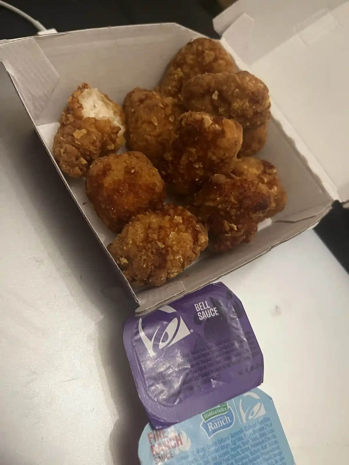 A white takeout box filled with golden-brown fried chicken nuggets sits on a white surface, with a purple packet labeled…
