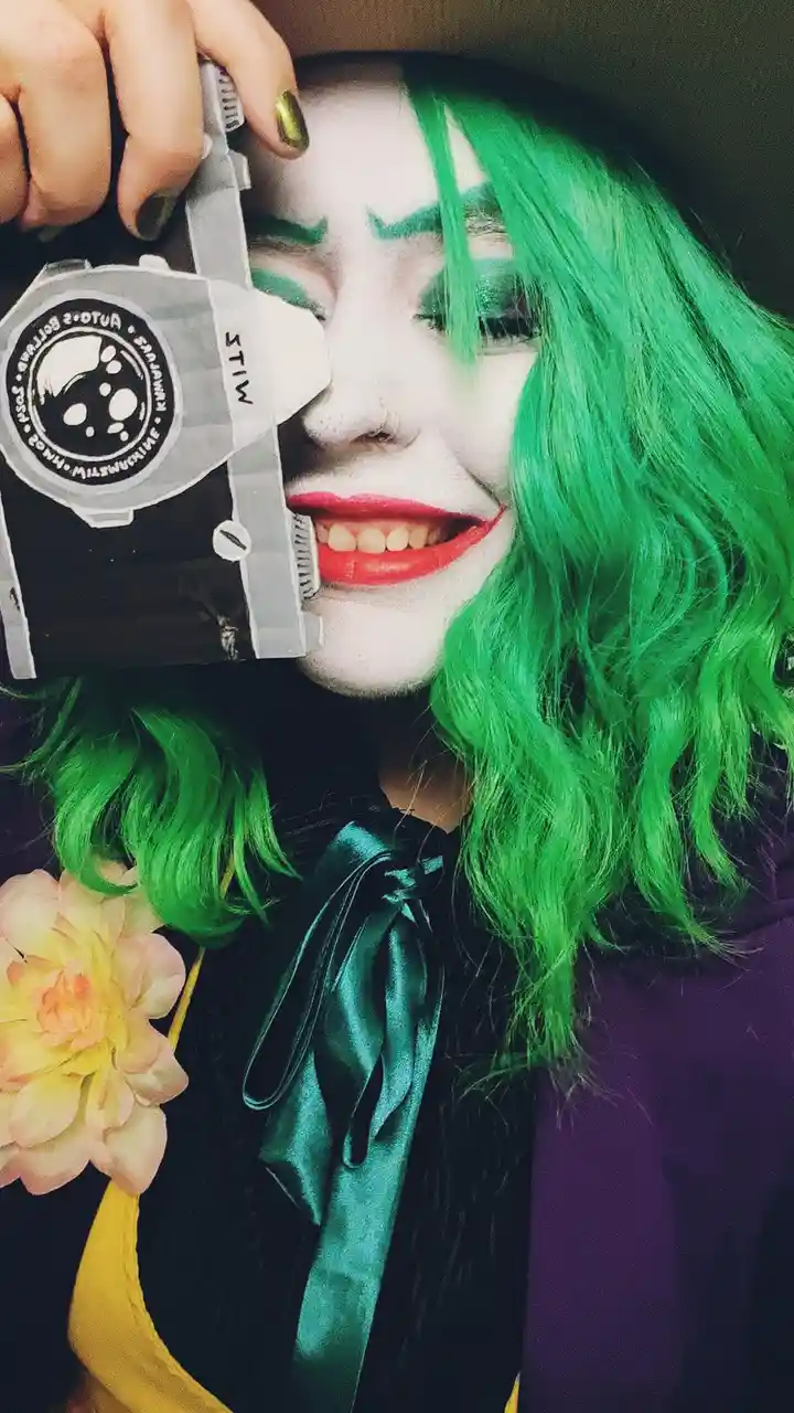 A person with bright green hair and white face paint, wearing red lipstick and green eye makeup, holds a homemade cardboard…
