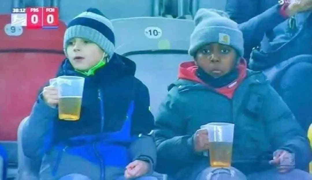 Two young boys in winter hats and jackets sit in stadium seats holding plastic cups of beer, looking intensely at the…