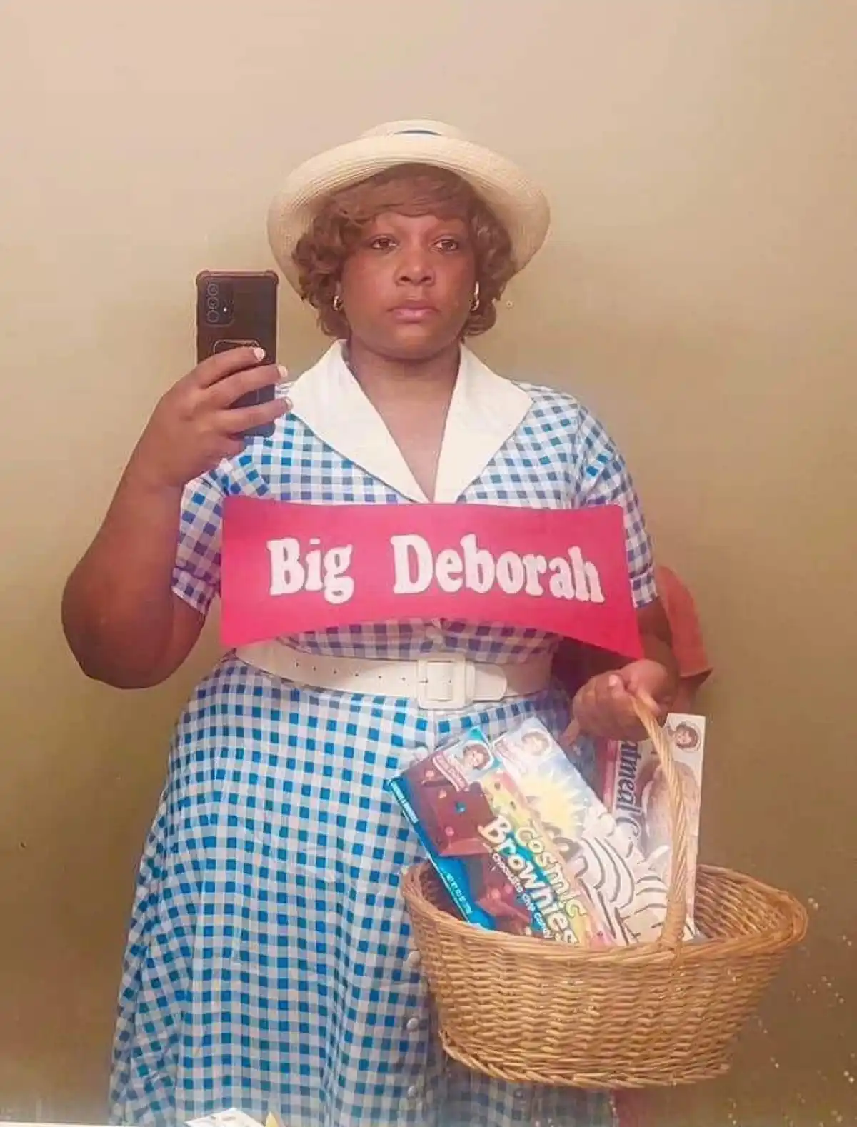 A person in a blue and white checkered dress with a white belt and straw hat takes a mirror selfie while holding a wicker…