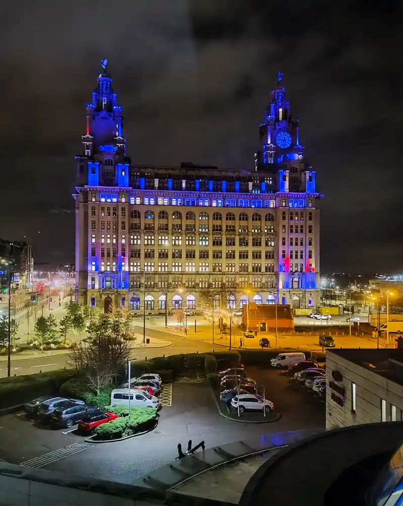 A nighttime photo of the iconic Liver Building in Liverpool, UK, illuminated with blue and red lights, with cars and…