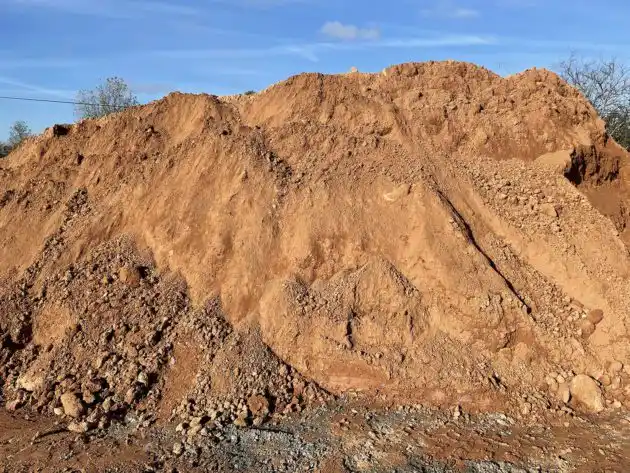 A large, uneven pile of reddish-brown dirt and gravel under a blue sky with sparse clouds. The image has text overlaid at…