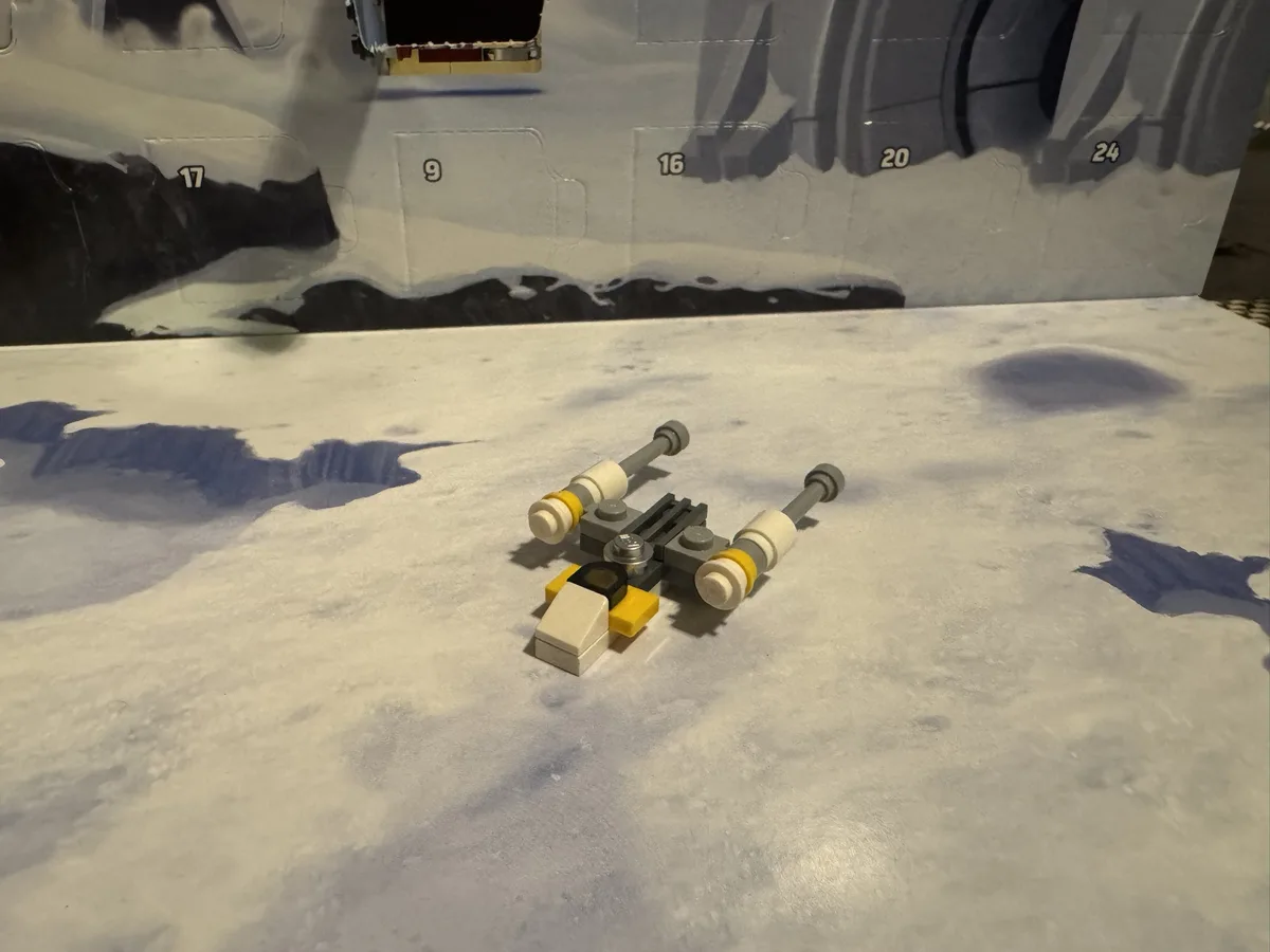 A small, crudely assembled Lego X-wing fighter model lies on a white, textured surface resembling a snowy or icy landscape…