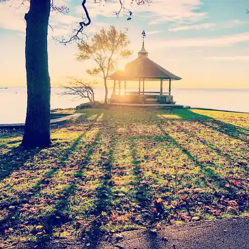 A serene autumn scene shows a wooden gazebo on a grassy hill overlooking water, with the sun setting behind it and casting…