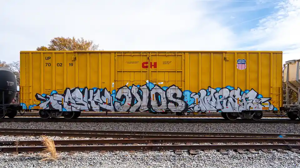 A yellow Union Pacific freight train car sits on tracks with large, stylized graffiti reading 'ALLUSIONS' painted across its…