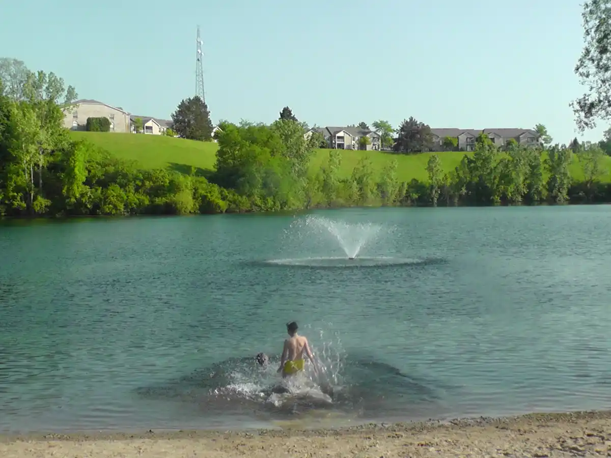 A shirtless person in yellow swim trunks splashes in a lake near a sandy shore, with a fountain spraying water in the middle…