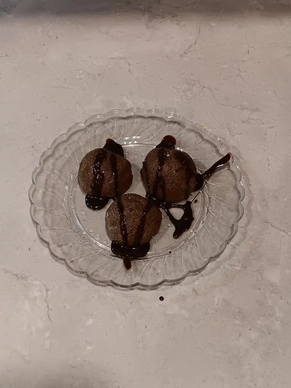Three round, brown, textured dessert balls sit on a clear glass plate with scalloped edges, drizzled with dark chocolate…