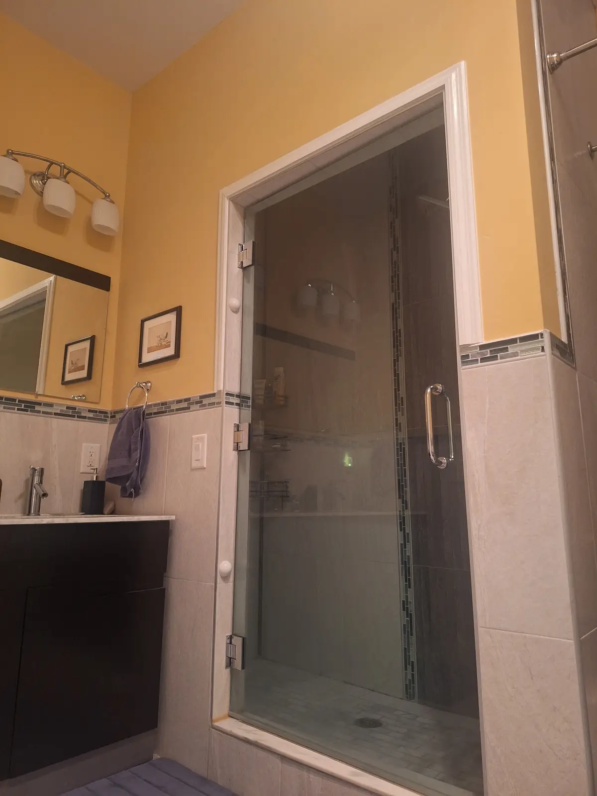 A bathroom with yellow walls, a tiled shower with a glass door, and a sink vanity. The glass shower door is visibly broken…