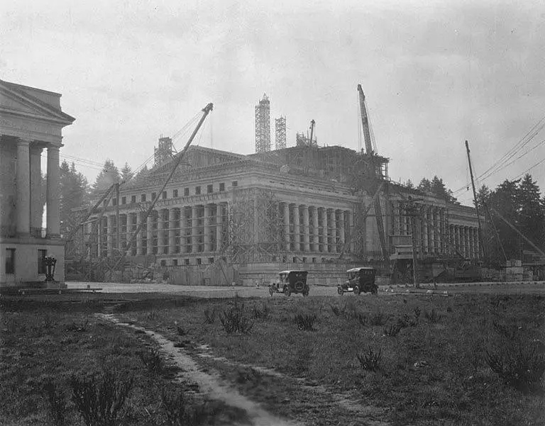 a black-and-white historical photo shows a large, ornate building under construction with cranes and scaffolding, surrounded…