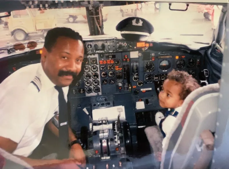 A vintage photo shows an adult Black man in a pilot uniform smiling at the camera from the cockpit of an airplane, while a…
