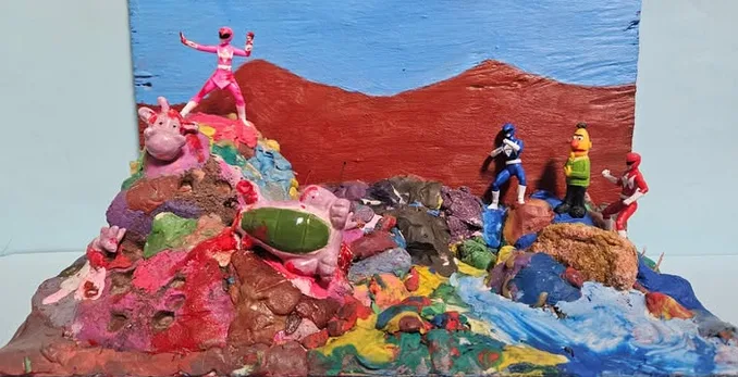 A handmade clay diorama depicts a scene with Power Rangers figurines. A pink Ranger stands triumphantly atop a colorful…