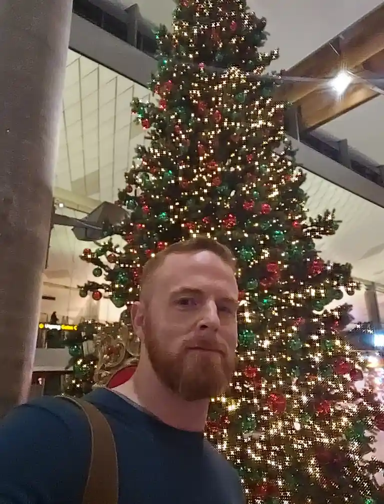 A man with a reddish beard and blue shirt takes a selfie in front of a large, brightly lit Christmas tree decorated with red…