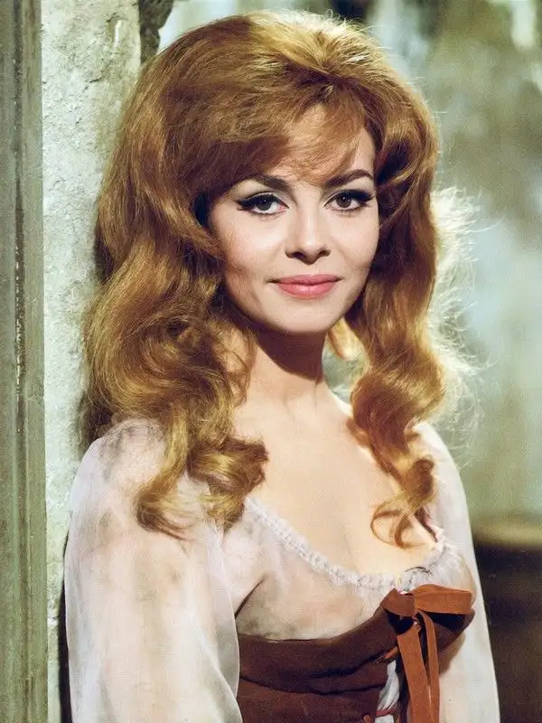 A vintage photograph of a woman with voluminous, wavy reddish-blonde hair and heavy eyeliner, wearing a sheer white blouse…