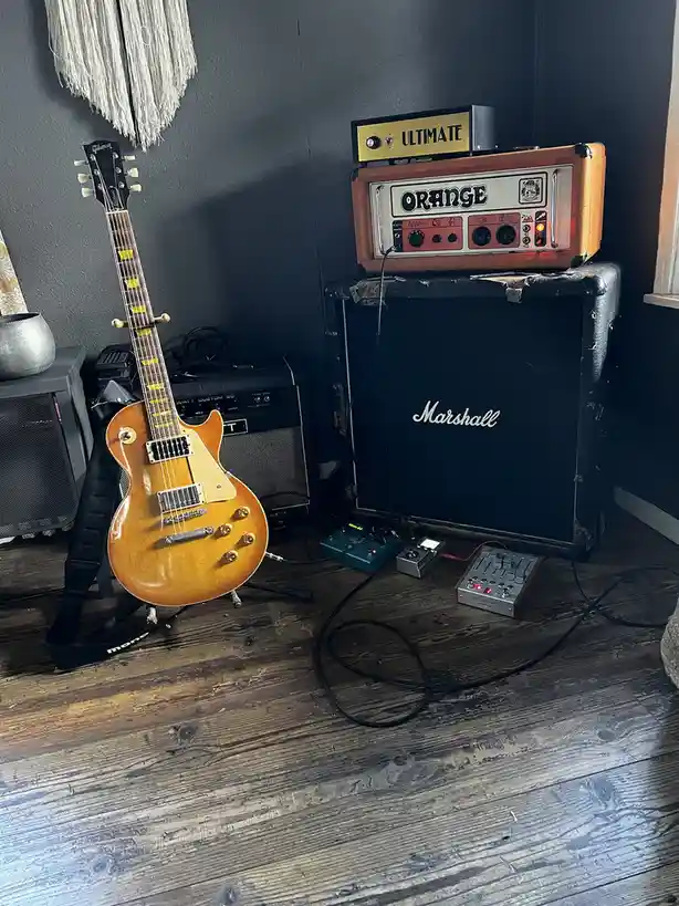 A Gibson electric guitar stands on a stand next to a stack of amplifiers, including an Orange amp head labeled 'ULTIMATE' on…