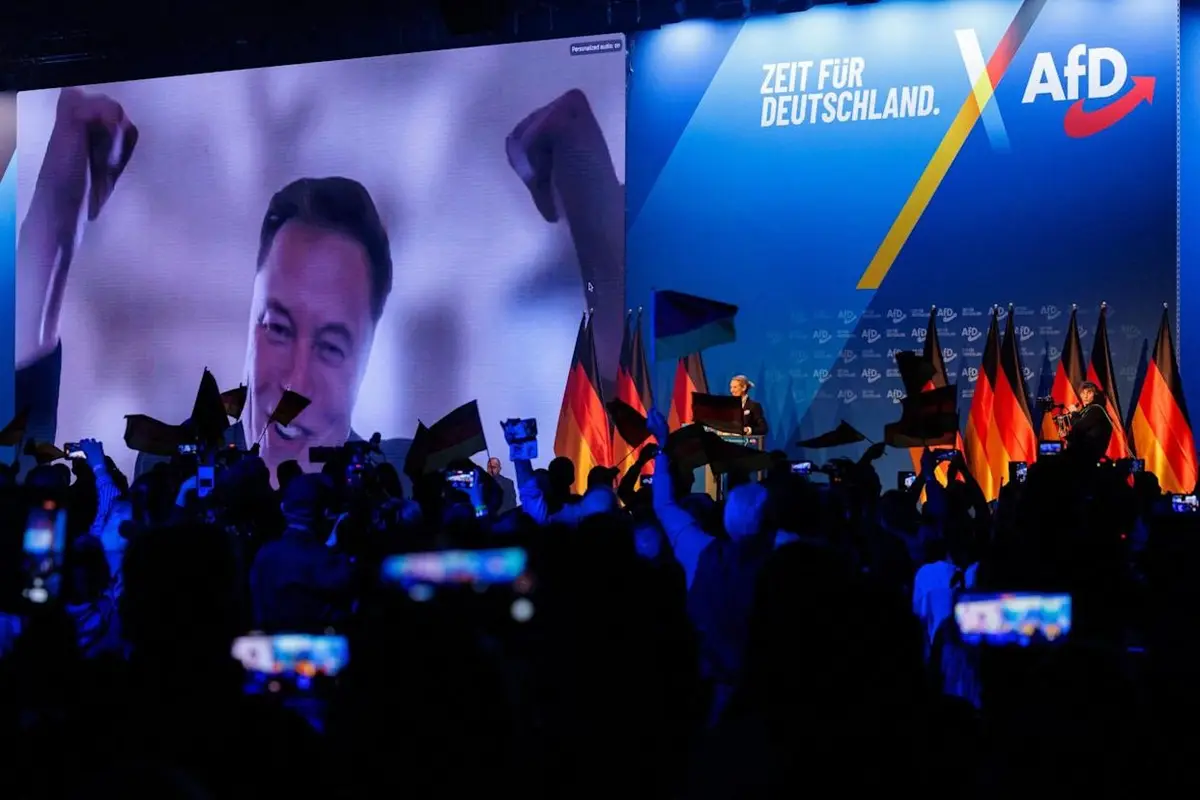 A large screen at an AfD political rally displays a smiling Elon Musk with raised fists, while a speaker stands at a podium…