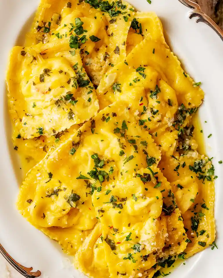 A close-up photo of a white platter filled with golden-yellow ravioli, garnished with chopped green herbs and grated cheese…
