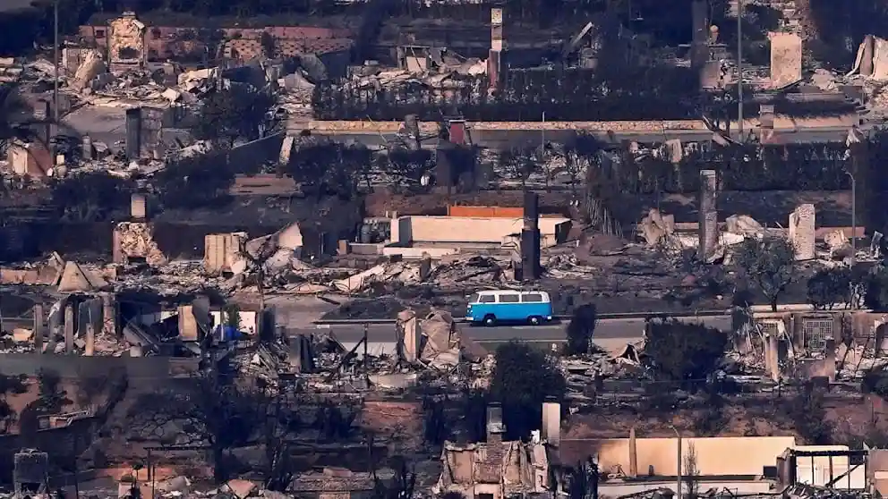 An aerial view shows a bright blue and white van driving down a paved road through a landscape of total destruction, with…