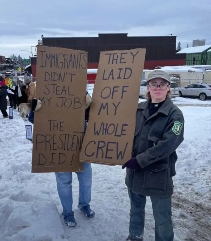 Two people stand in snow holding cardboard signs. The person on the left holds a sign reading 'IMMIGRANTS DIDN'T STEAL MY…