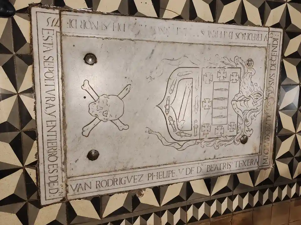 A close-up of a weathered, white marble tombstone embedded in a black-and-white geometric tiled wall. The stone features…