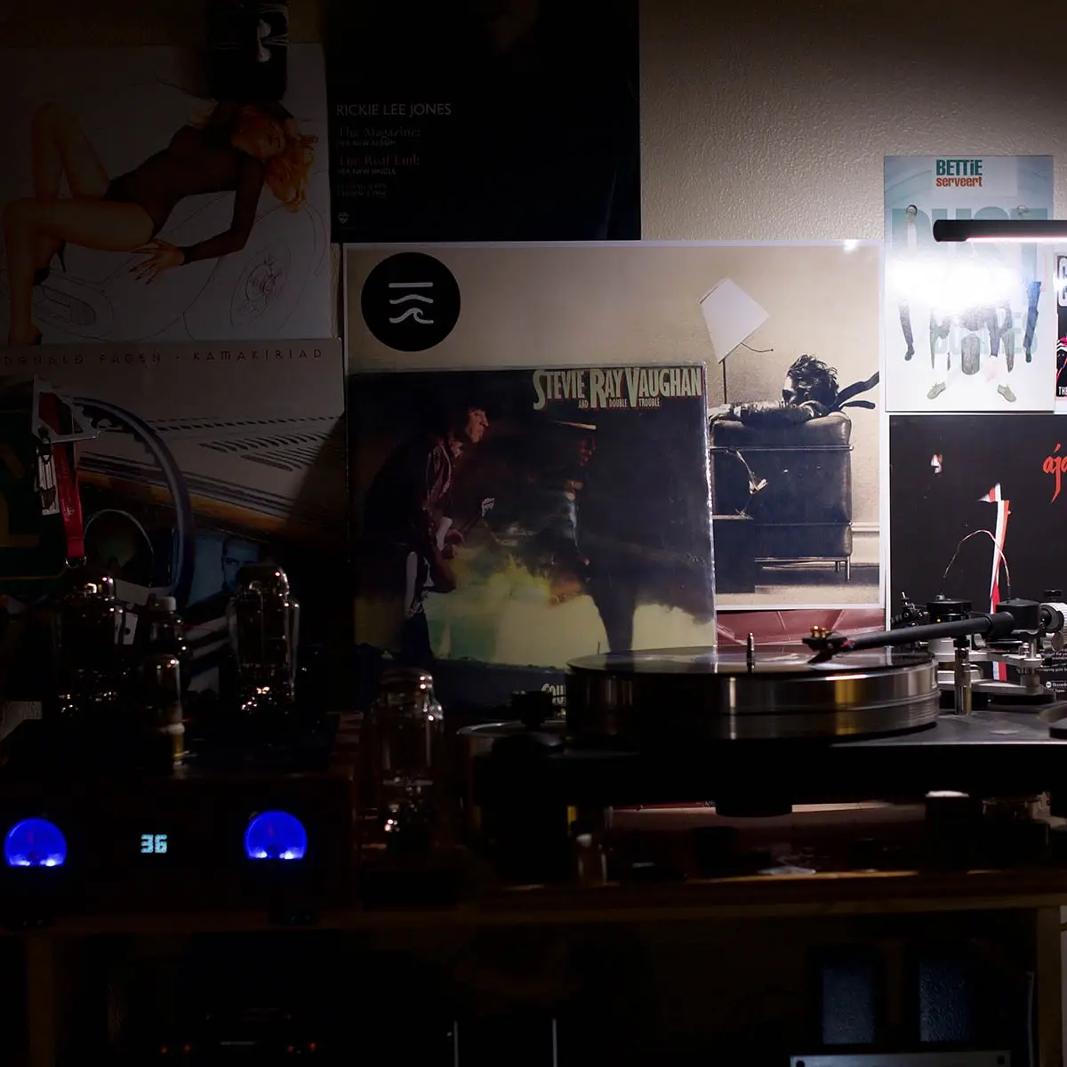 A dimly lit room features a turntable with a Stevie Ray Vaughan album cover standing upright beside it. The album art shows…