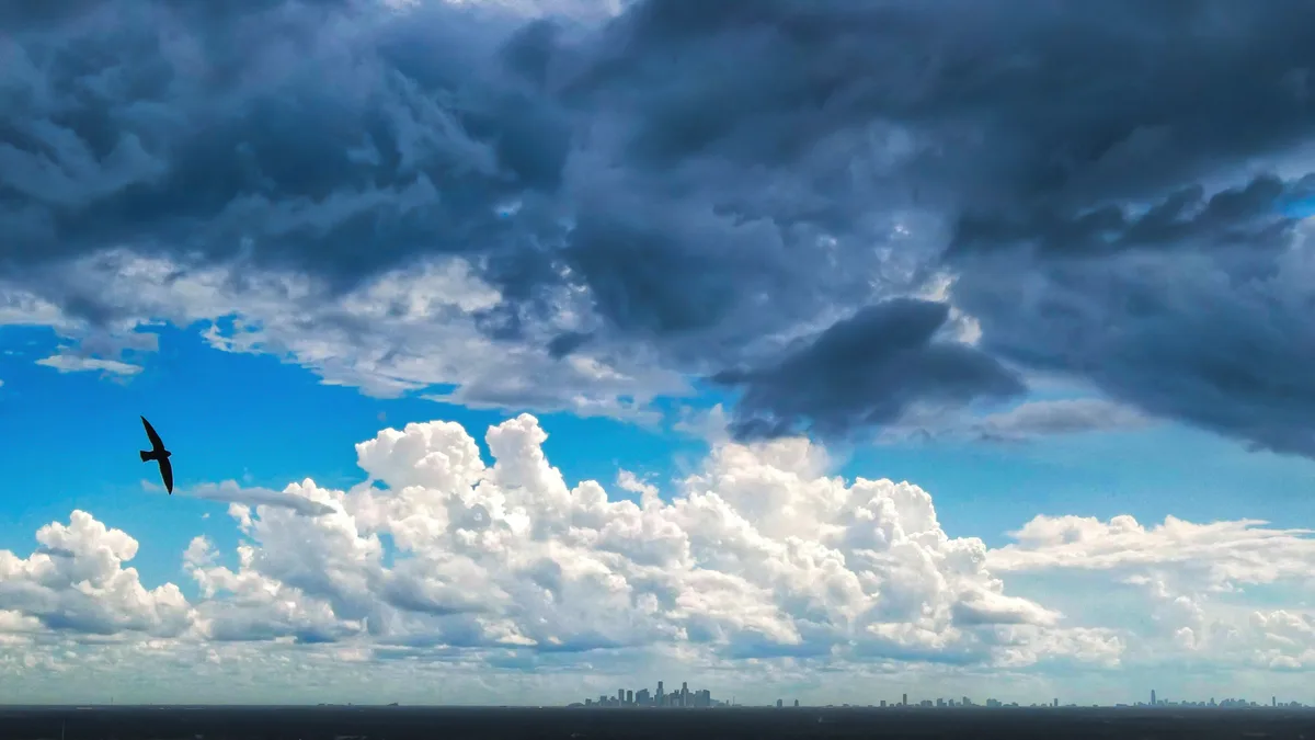 A dramatic sky with dark storm clouds overhead and bright white cumulus clouds below, with a distant city skyline on the…